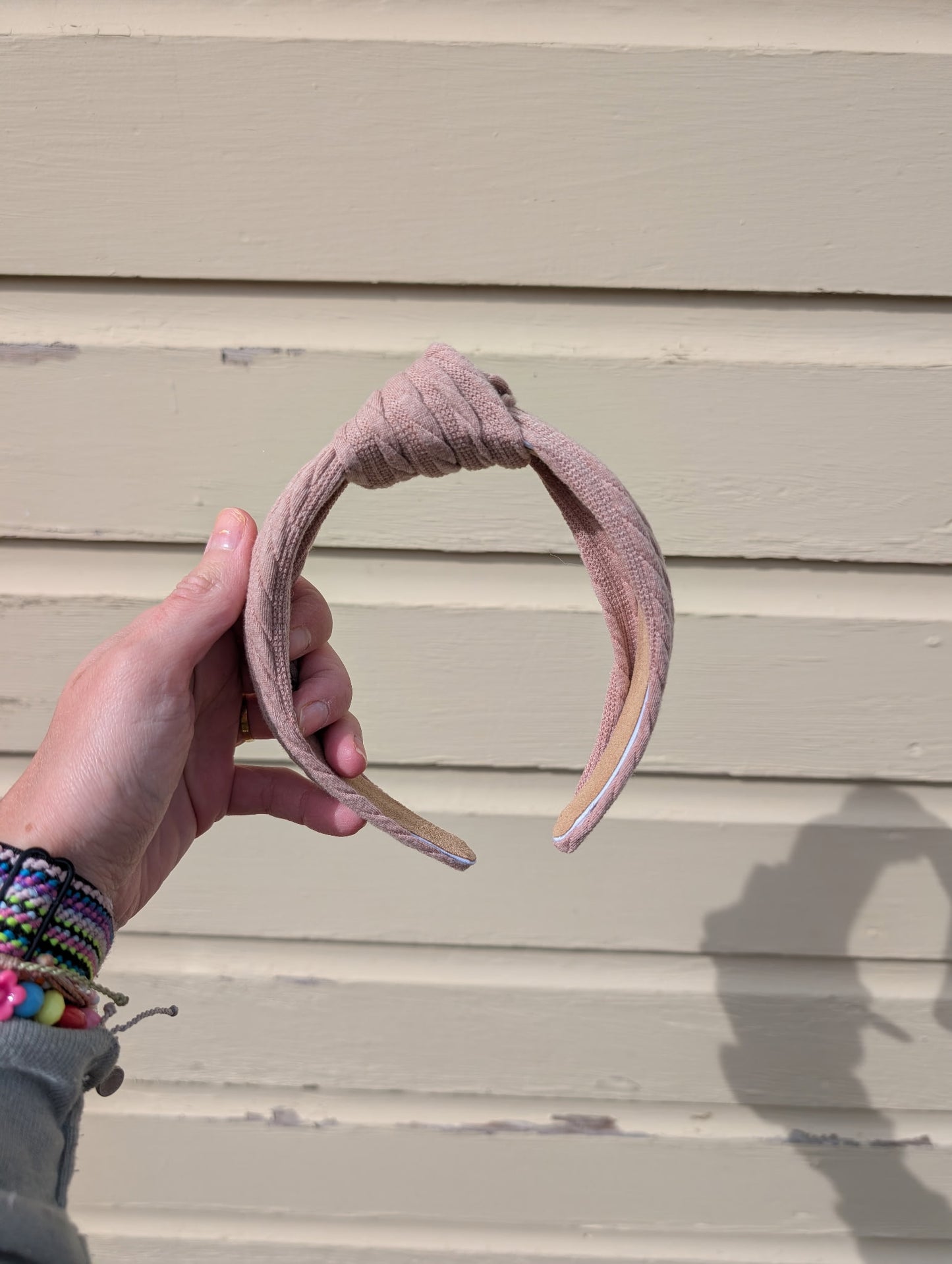 Hand holding a taupe headband against a beige wall.