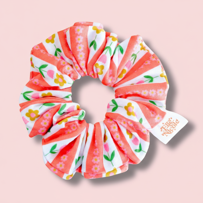 A hair scrunchie featuring a white background with vertical coral-pink stripes accented by tiny pink and yellow tulip illustrations, finished with a branded fabric tag.
