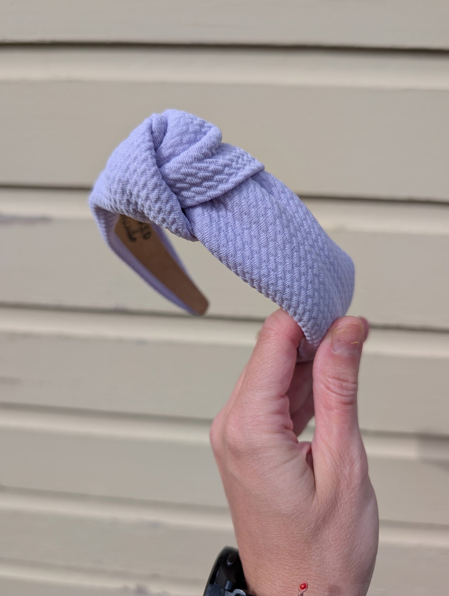 Hand holding a light purple textured headband against a beige wall.