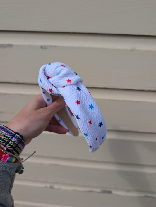 Hand holding a knotted white headband with colorful  red, white, and blue stars against a beige wall.