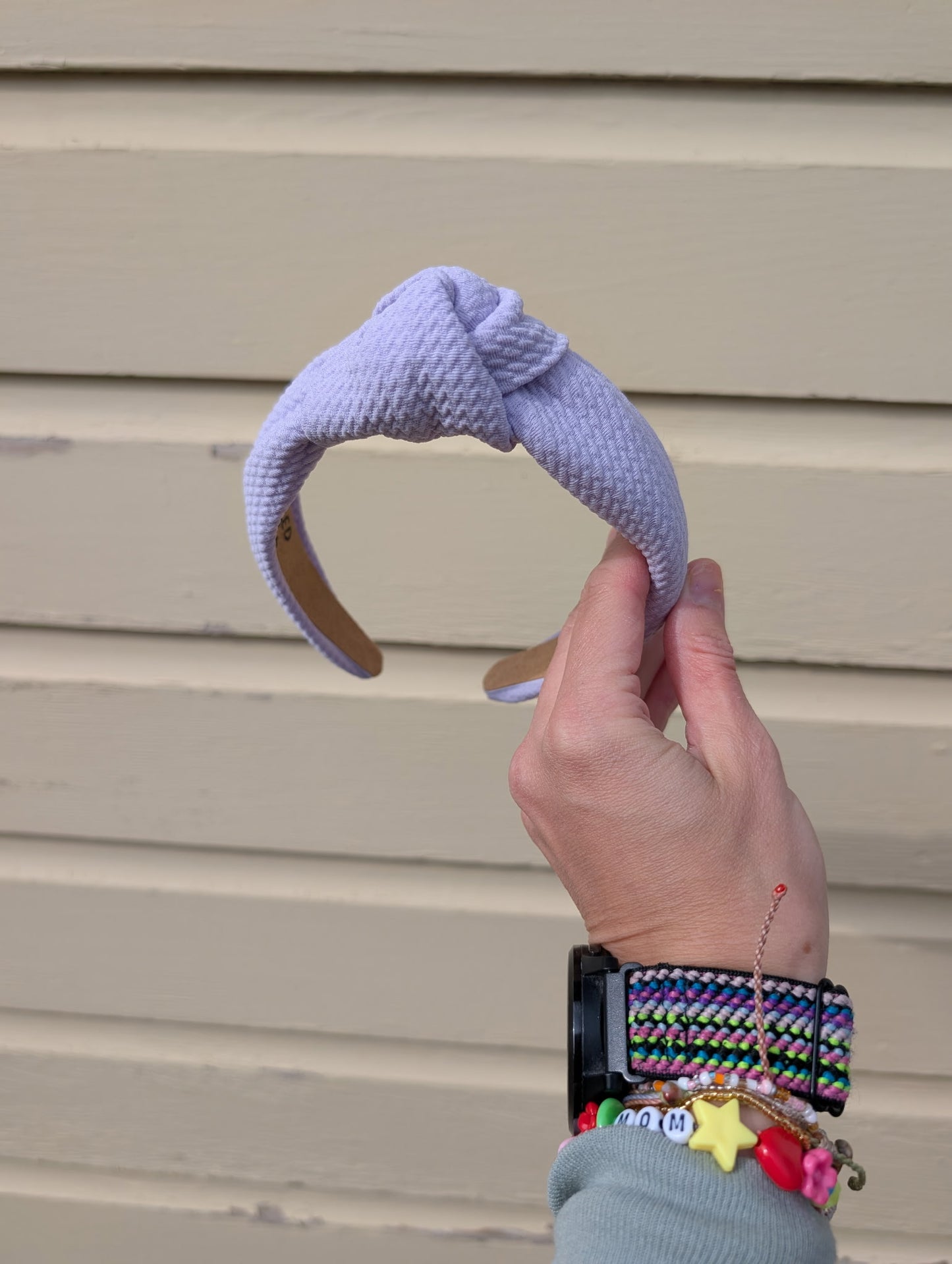 Hand holding a light purple knitted headband against a beige wall.