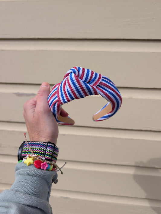 Hand holding a colorful blue and red striped headband against a beige wall.