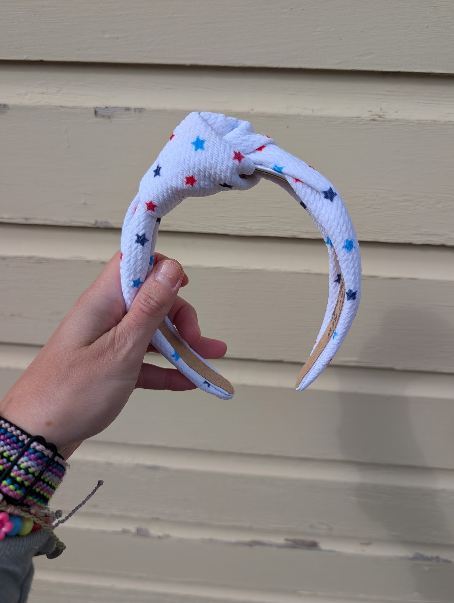 Hand holding a knotted white headband with colorful  red, white, and blue stars against a beige wall.