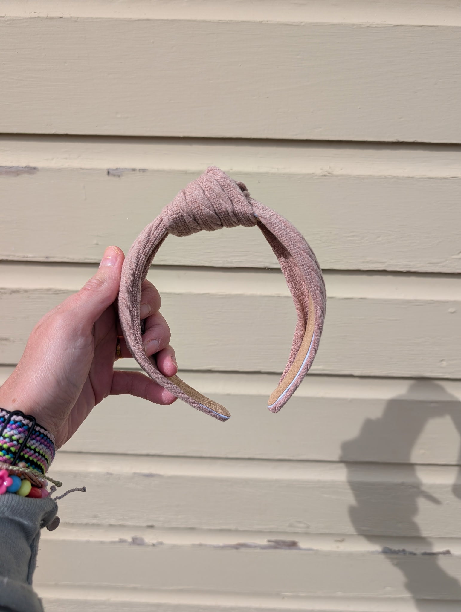 Hand holding a taupe headband against a beige wall. 