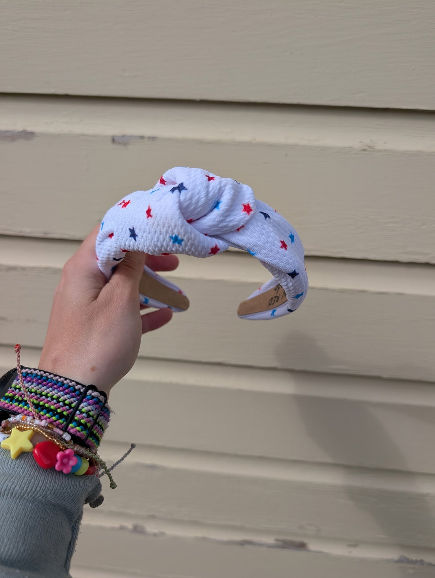 Hand holding a knotted white headband with colorful  red, white, and blue stars against a beige wall.