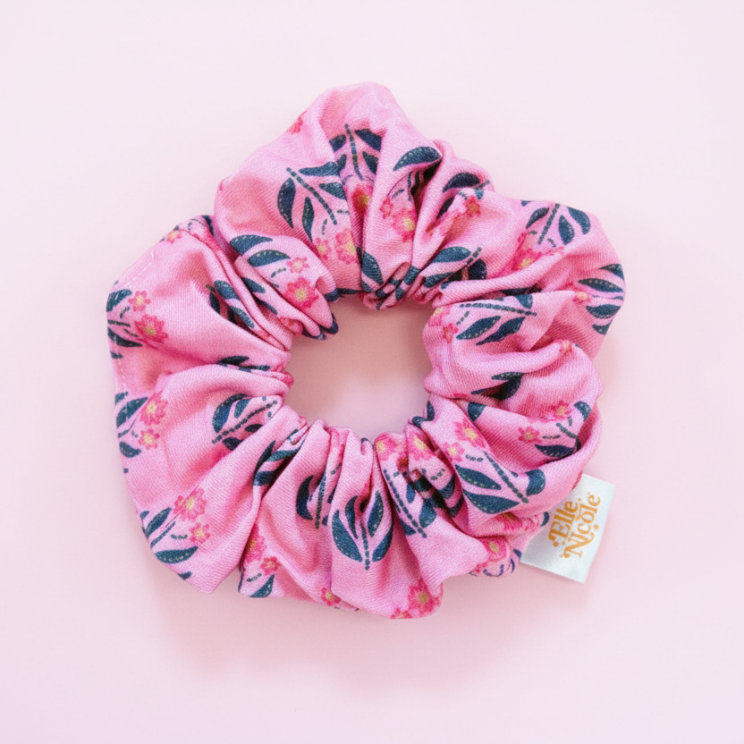 A soft pink hair scrunchie featuring a delicate botanical floral print with muted green leaves and small pink flowers, finished with a branded fabric tag.