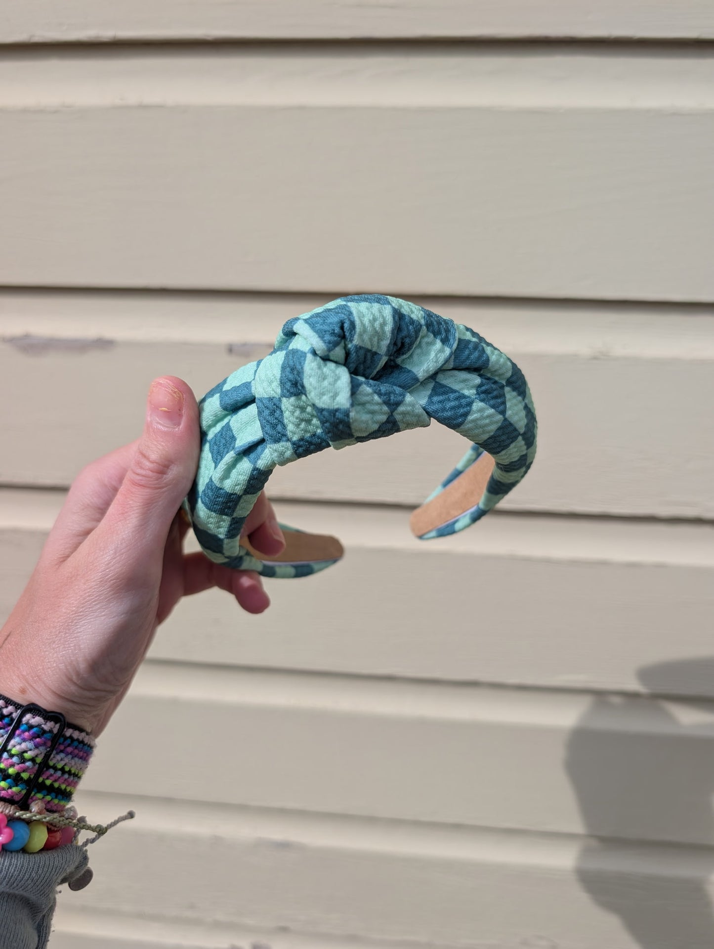 Hand holding a teal and green checkered headband against a beige wall. 