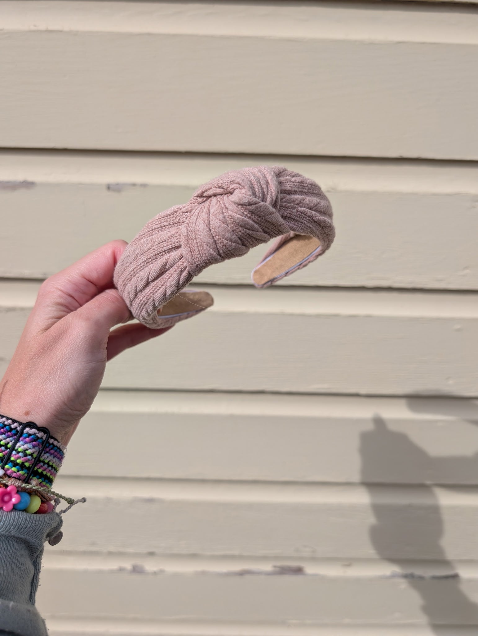 Hand holding a taupe headband against a beige wall. 