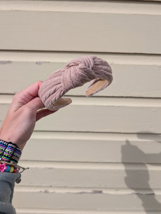 Hand holding a taupe headband against a beige wall. 
