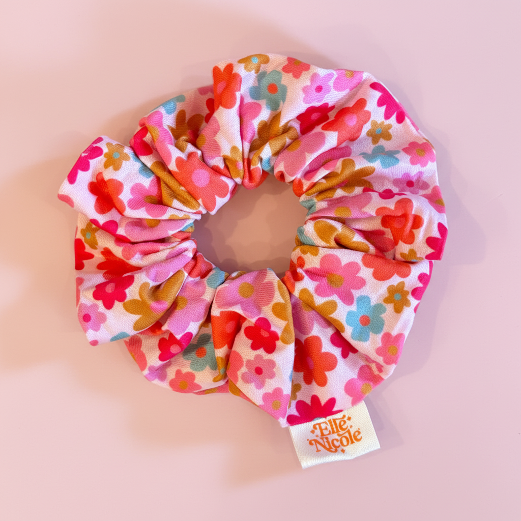 A colorful hair scrunchie featuring a busy 70s-inspired floral pattern with shades of pink, orange, and blue on a light background, finished with a branded fabric tag.