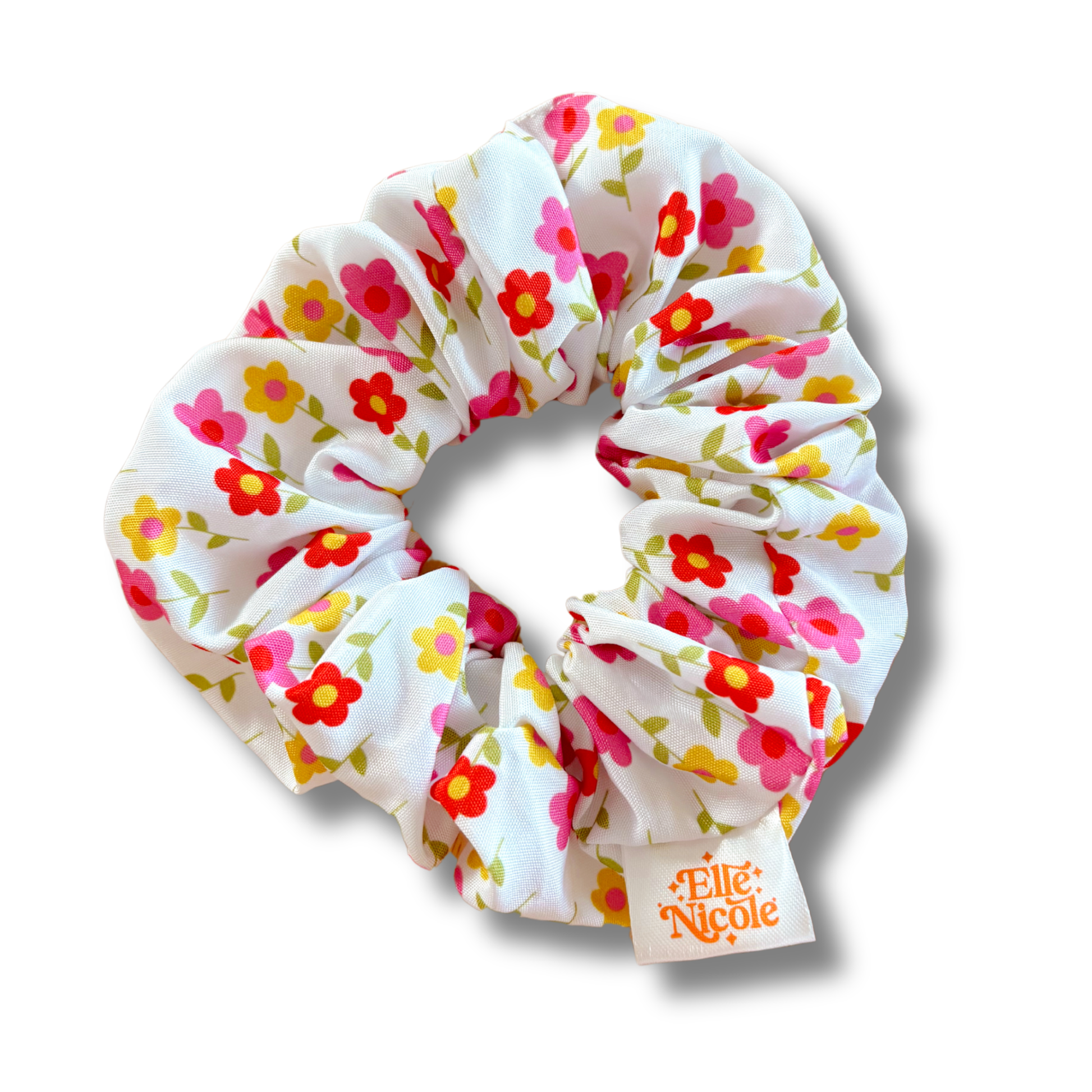 A white hair scrunchie featuring a whimsical pattern of dainty flowers in pink, yellow, and red with green stems, finished with a branded fabric tag.