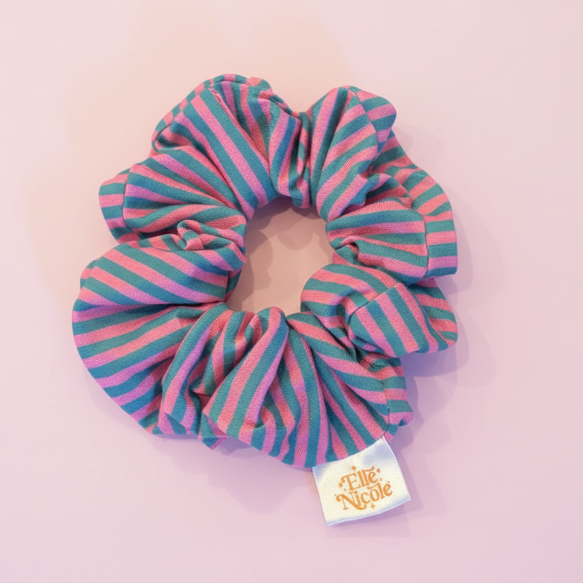 A hair scrunchie with a classic, candy-colored alternating stripe pattern in muted pink and blue tones, finished with a branded fabric tag