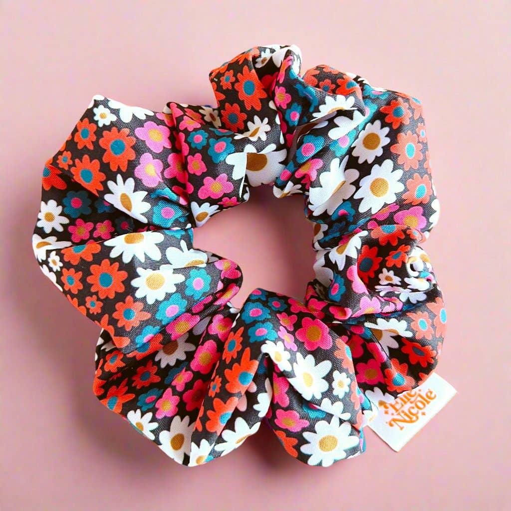 A high-contrast hair scrunchie with a dark background and a vibrant floral pattern in bright pink, orange, and blue, finished with a branded fabric tag