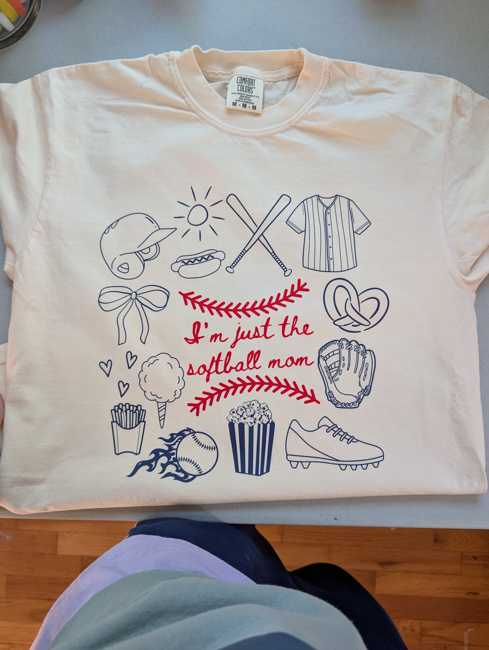 A cream-colored Comfort Colors t-shirt featuring blue and red doodles of baseball icons like bats, a glove, popcorn, and a pretzel, with central text that reads "I'm just the softball mom."