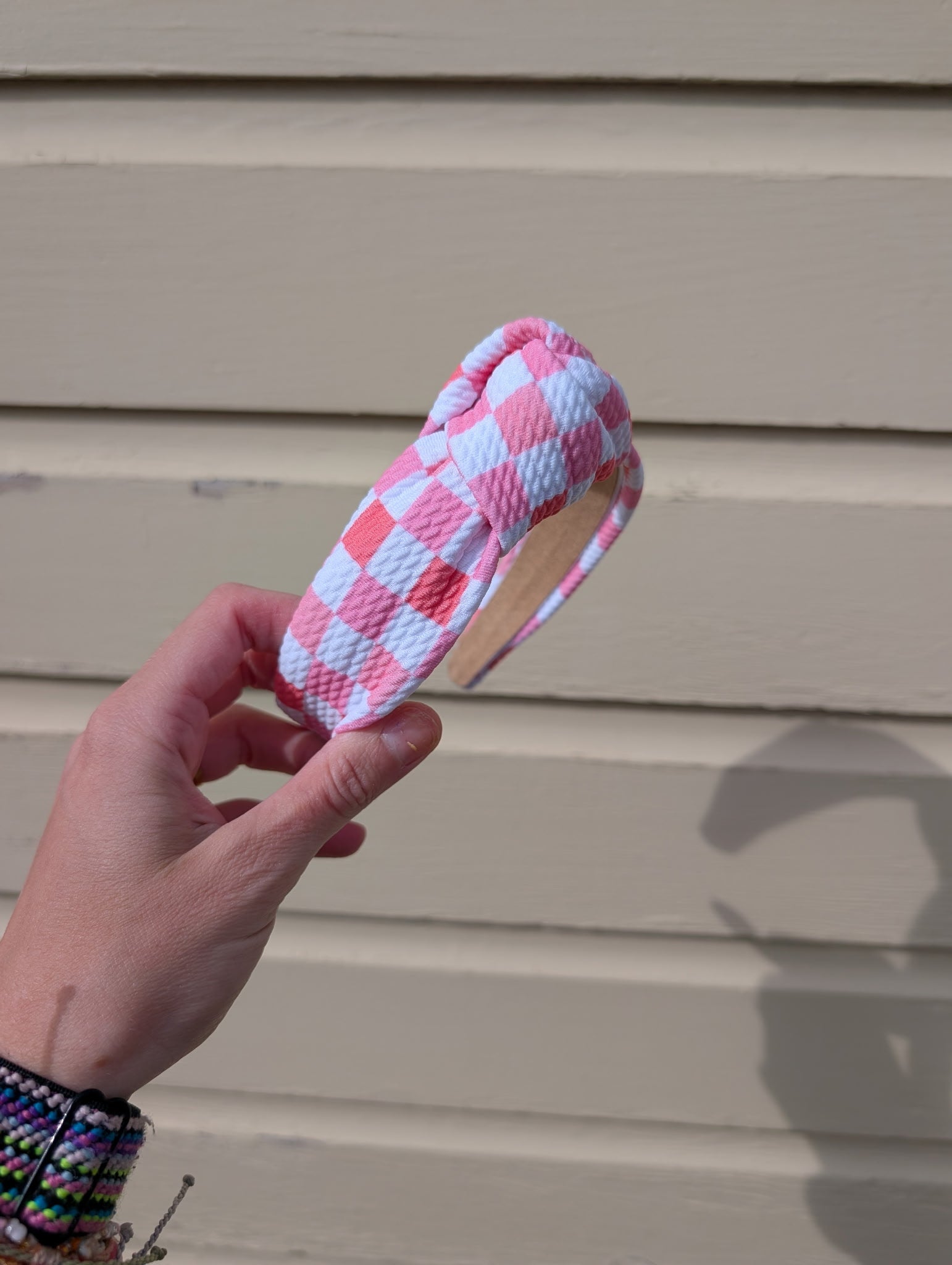 Hand holding a pink and white checkered headband against a beige wall.