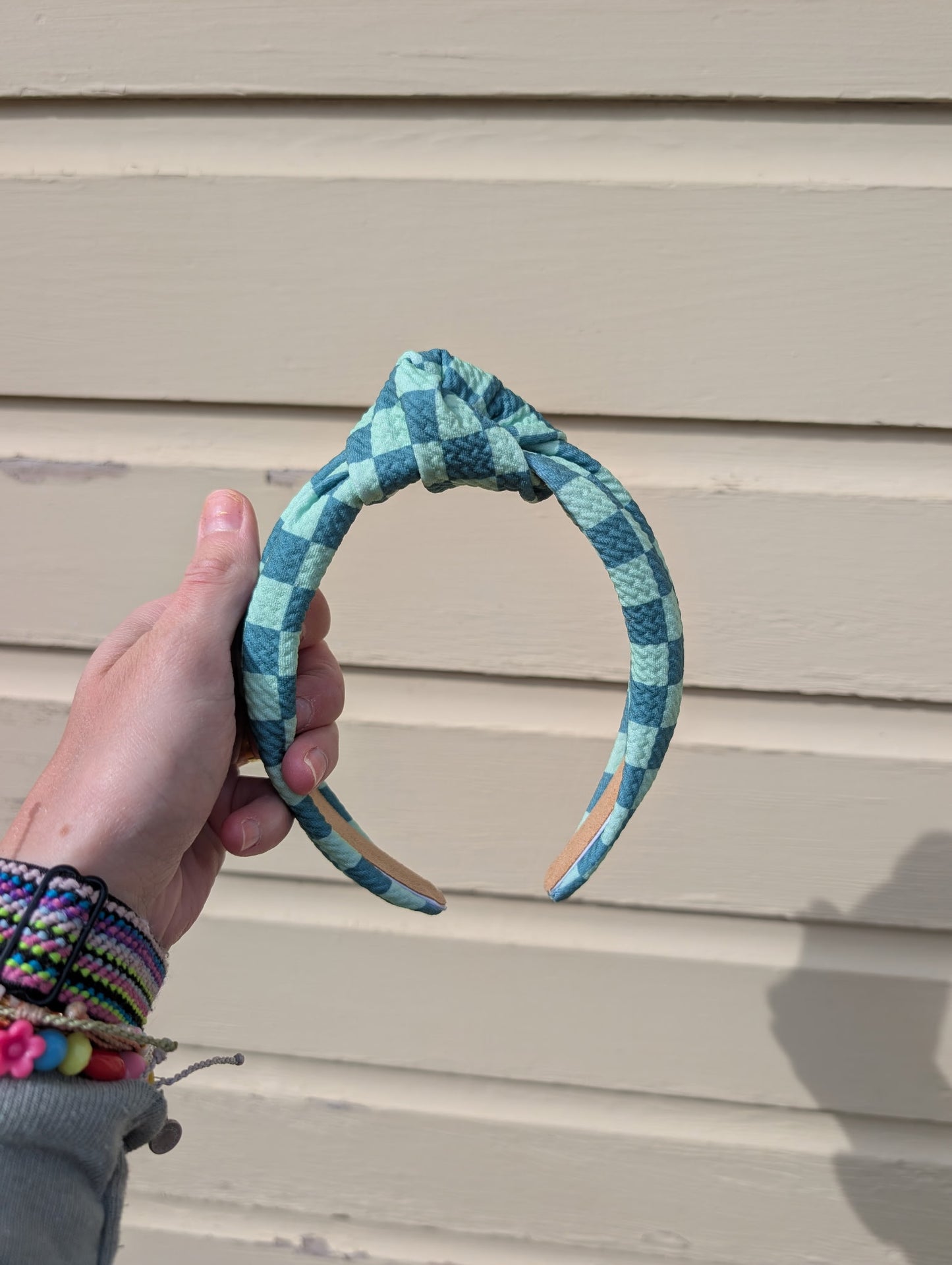 Hand holding a teal and green checkered headband against a beige wall. 